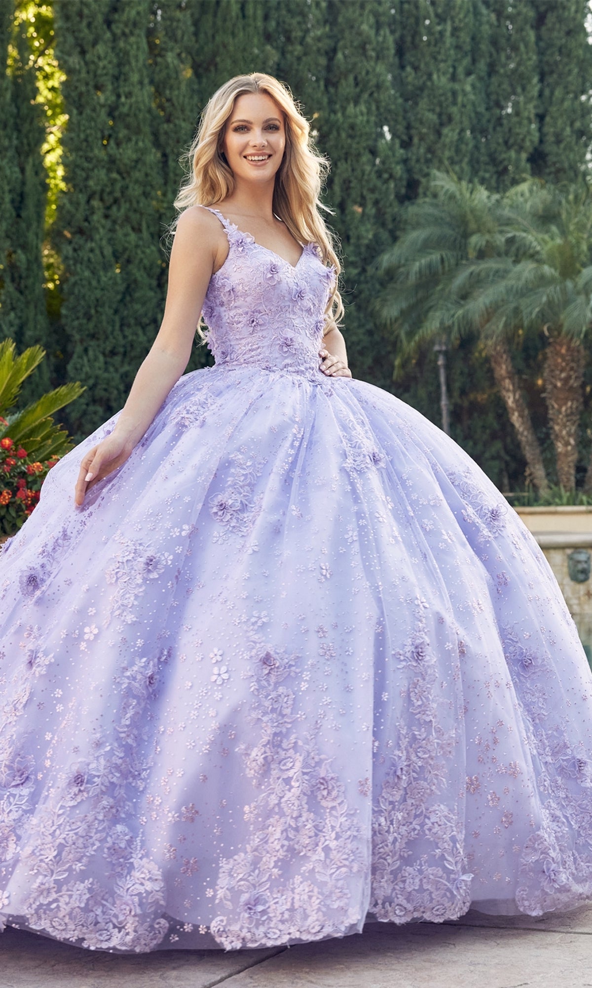 A young woman stands outdoors in the Juliet 1446 Lilac Purple Quinceañera Dress with floral embellishments, surrounded by trees and greenery.